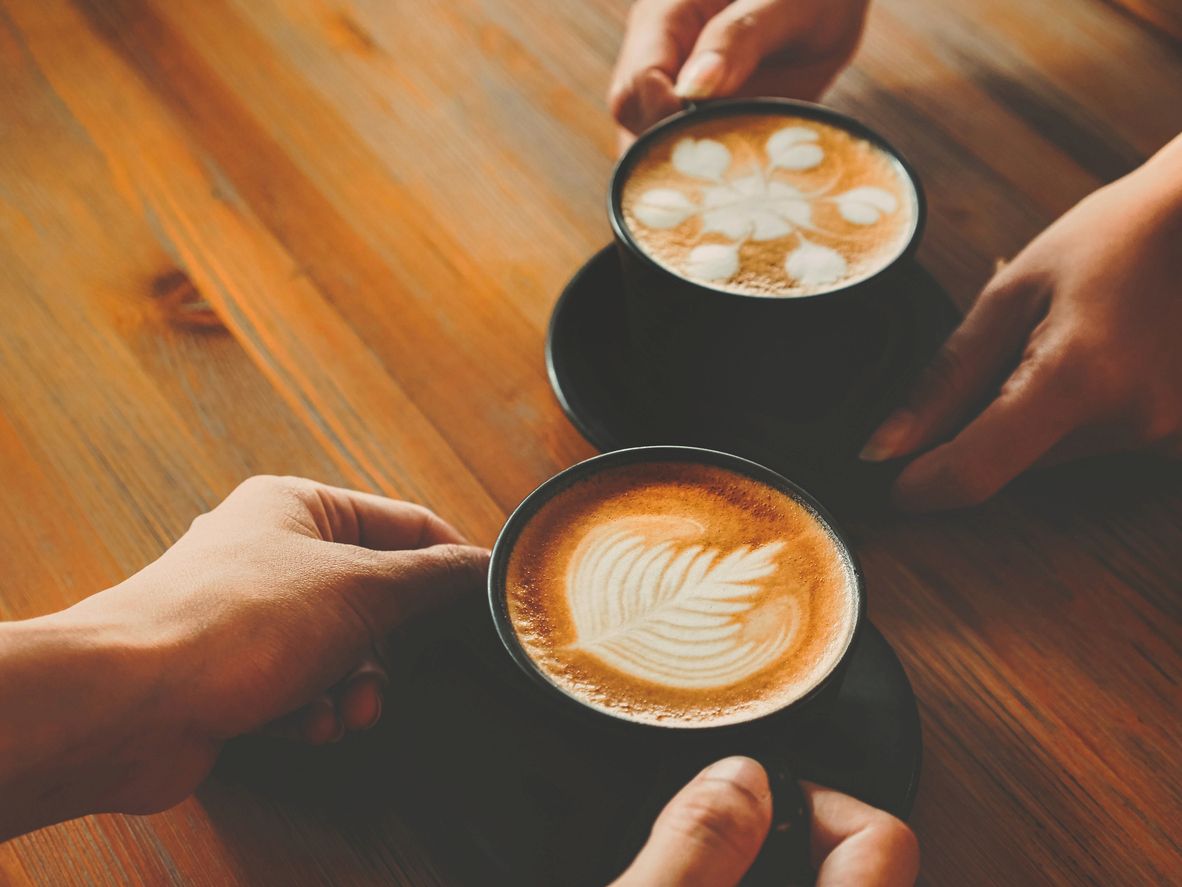 Two lattes on a brown wooden table with two pairs of hands holding by the handle and saucer.