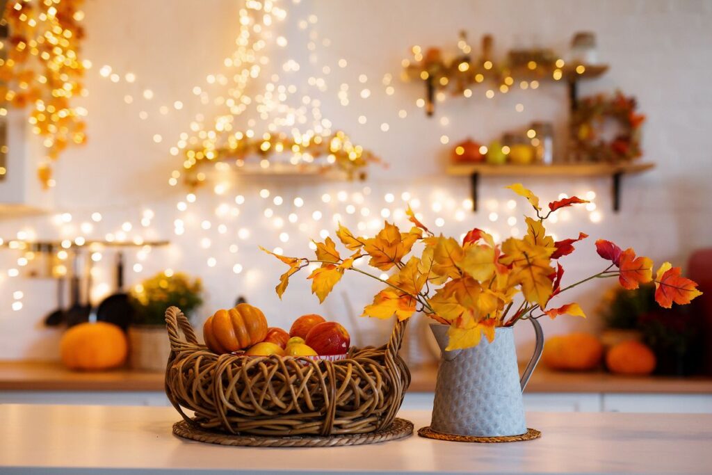 A kitchen counter with a cornucopia with pumpkins next to a pitcher of golden leaves with twinkling lights behind.