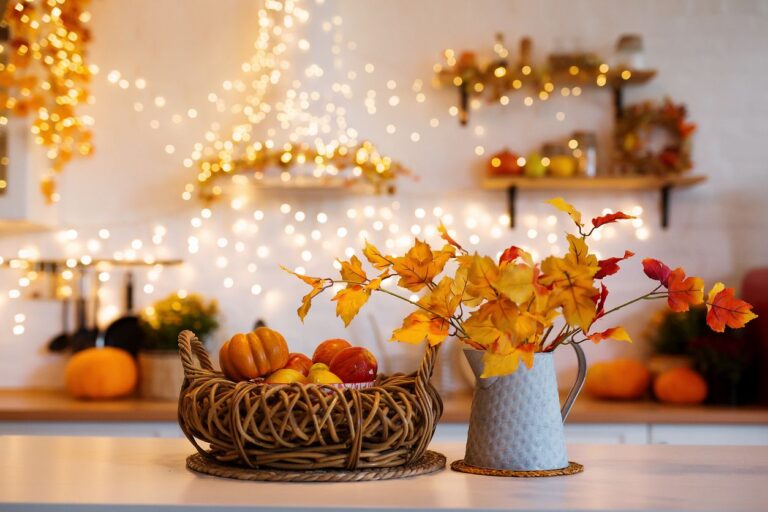 A kitchen counter with a cornucopia with pumpkins next to a pitcher of golden leaves with twinkling lights behind.