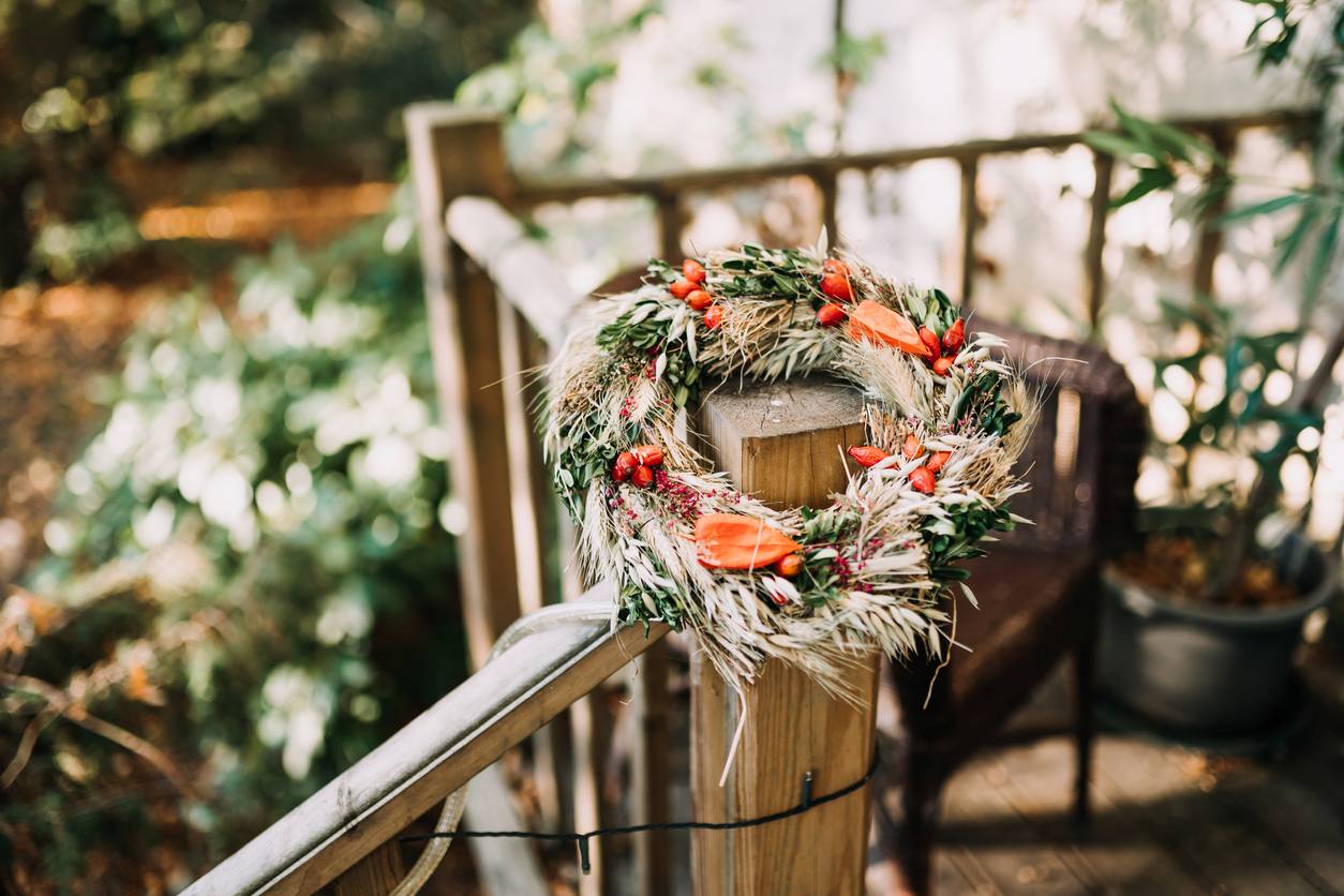A wreath made of dried plants and goldenrain tree seeds, wrapped in evergreen and wheat, sits on a deck railing. 