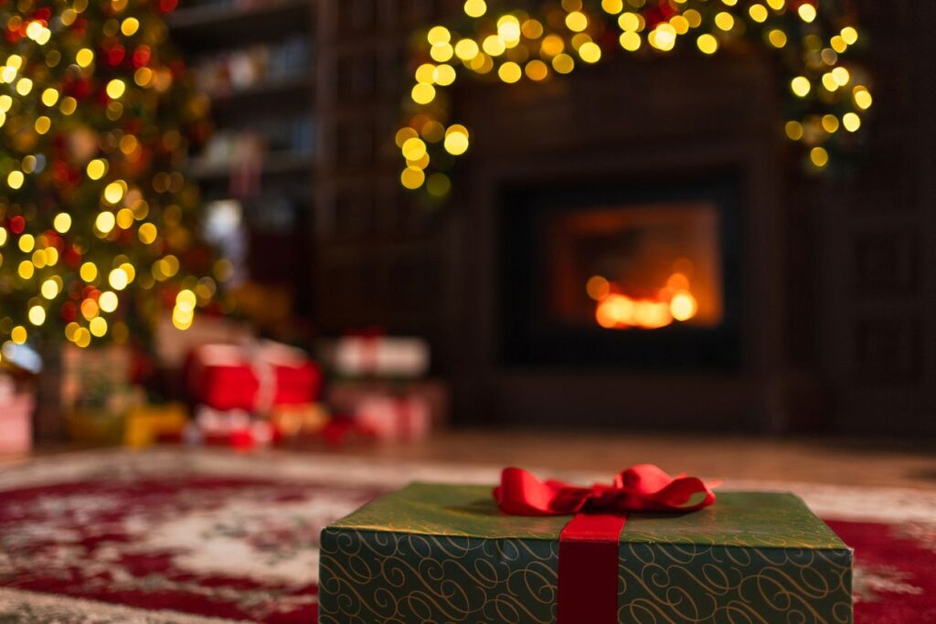 A gift wrapped in green paper and a red bow sits by a Christmas tree and a lit fireplace decorated with lights.